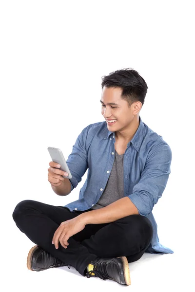 Junger asiatischer Mann schaut auf das Handy, während er auf dem Boden sitzt — Stockfoto