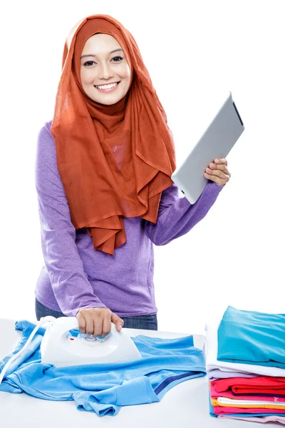 Young woman wearing hijab reading article on tablet while ironin — Zdjęcie stockowe