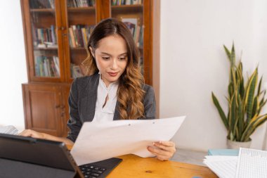 Asyalı kadın analist, masa başında doküman ve raporları incelerken, yoğunlaştırılmış ifade, rakamları ve tableti kontrol ederken, düzenli