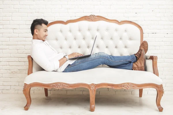 Азіатський молодий чоловік сидить розслабитися на дивані під час роботи на ноутбуці — стокове фото