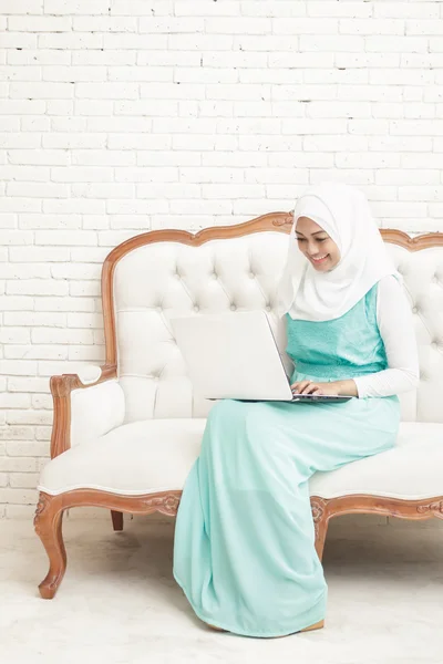 Wanita muda Asia tersenyum mengenakan jilbab duduk di sofa sementara wo — Stok Foto