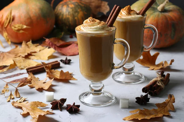 Two glass cups pumpkin latte with spices on grey background with pumpkins and autumn leaves, close-up.