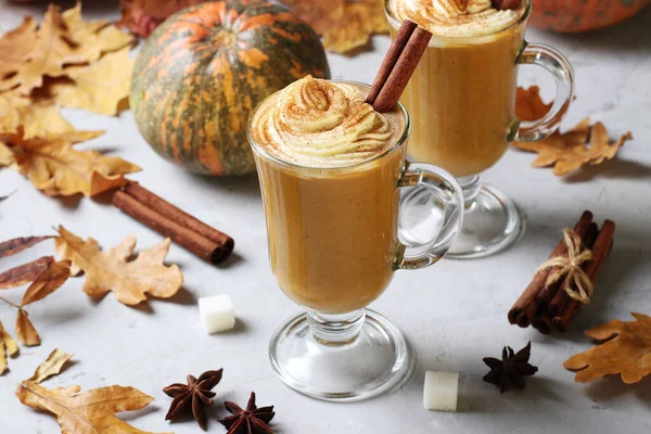 Two glass cups pumpkin latte with spices and creamy foam on grey background with pumpkins and autumn leaves.