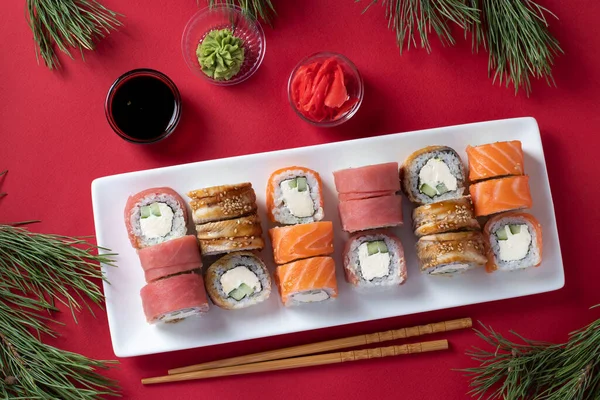 Świąteczna kolacja z łososiem, tuńczykiem i sushi z filadelfijskim serem na białym talerzu na czerwonym tle. Podawany z sosem sojowym, wasabi, marynowanym imbirem i kijami do sushi. Widok z góry — Zdjęcie stockowe