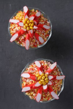 İki porsiyon yengeç çubuklu salata, koyu arkaplanda şeffaf cam kaselerde yumurta ve mısır. Üst görünüm