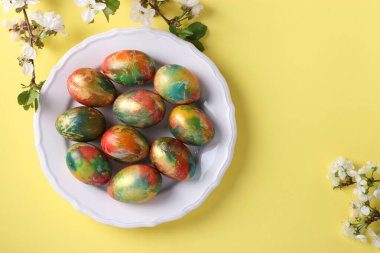 Paskalya için besin renklerine boyanmış mermer yumurtalar sarı arka planda beyaz bir plaka üzerine yerleştirilmiş. Yukarıdan görüntüle.
