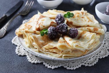 Buğday unu, yumurta ve kefir ile ince krepler, böğürtlen, reçel ve gri masada bir bardak süt ile servis edilir.