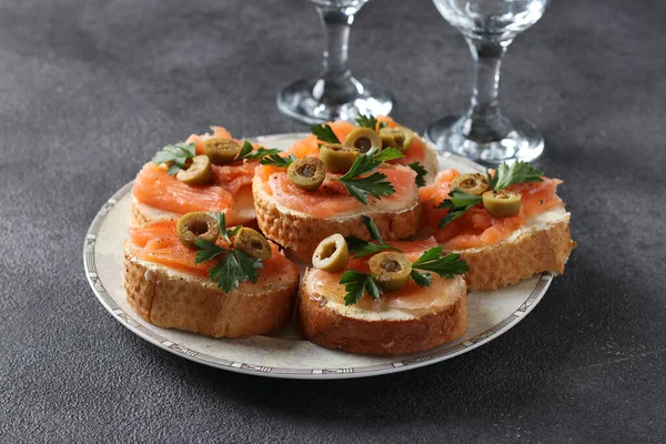 Палички солоного лосося, зелених оливок і петрушки на грінках білого хліба на темно-сірому фоні. Святкова закуска . — стокове фото
