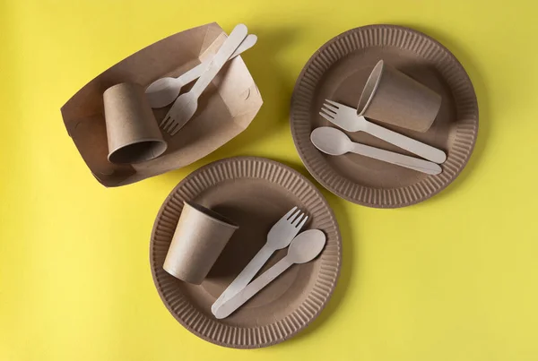 Three sets disposable biodegradable tableware of craft paper and wood - plates, forks, spoons, glasses on yellow background. Zero Waste. View from above.