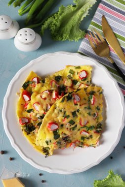 Açık mavi arka planda beyaz tabakta yengeç çubuklu, peynirli ve yeşil soğanlı omlet. Üst görünüm