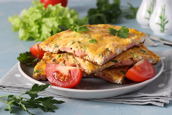 Omelet med pølser, ost og persille serveret med tomater på en grå plade på en lyseblå baggrund. Nærbillede. - Stock-foto