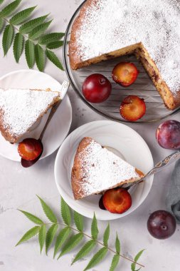 Ev yapımı erikli turta, arka planda tabaklarda kesik parçalar var. Pastaya pudra şekeri serpilmiş. Üst görünüm