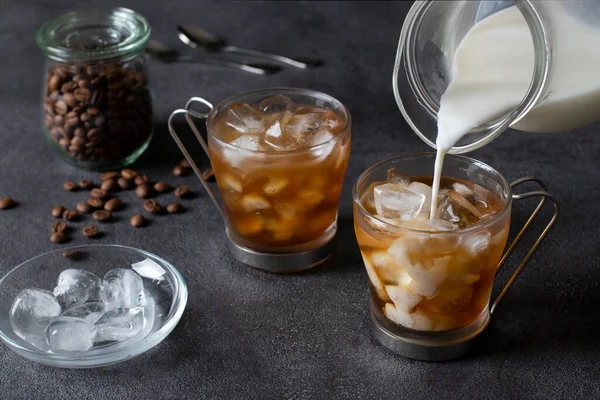 stock image Two cups of cold coffee with ice cubes, into which milk is being poured from a jug. Dark background. Close-up.