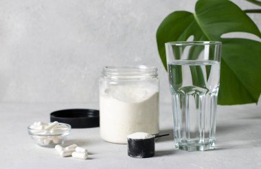 Collagen powder in a jar and measuring spoon, collagen capsules and a glass of water on a gray background with monstera leaf. Healthy and antiage concept