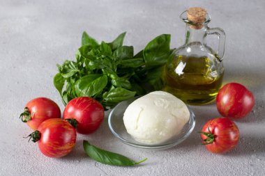 Caprese İtalyan salatası yapmak için gerekenler: domates, mozzarella peyniri, fesleğen ve açık gri arka planda zeytinyağı