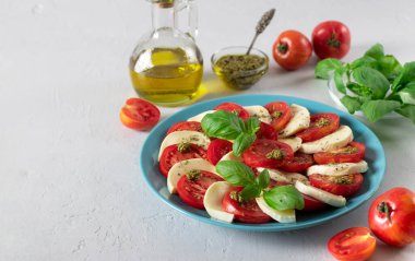Dilimlenmiş domatesli İtalyan caprese salatası, mozzarella peyniri, fesleğen ve açık gri arka planda mavi tabakta zeytinyağı. Boşluğu kopyala