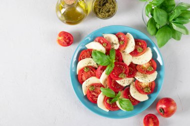 Dilimlenmiş domatesli İtalyan caprese salatası, mozzarella peyniri, fesleğen ve açık gri arka planda mavi tabakta zeytinyağı. Uzayı kopyala Üst görünüm