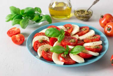 Dilimlenmiş domatesli İtalyan caprese salatası, mozzarella peyniri, fesleğen ve açık gri arka planda mavi bir tabakta zeytinyağı..