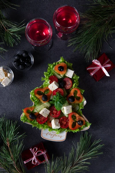 Gesunder griechischer Salat auf Teller serviert als Weihnachtsbaum mit festlicher Dekoration und zwei Gläsern Wein auf dunklem Hintergrund. Vertikales Format. Ansicht von oben — Stockfoto