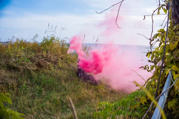 A boy in a black sports suit sits on the green grass, the boy has a crimson smoke bomb in his hand and it smokes into the sky.