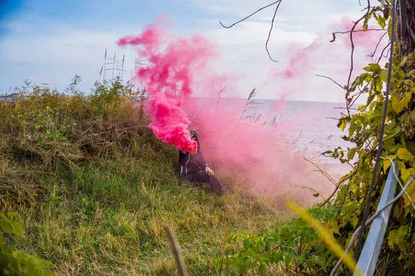 A boy in a black sports suit sits on the green grass, the boy has a crimson smoke bomb in his hand and it smokes into the sky.