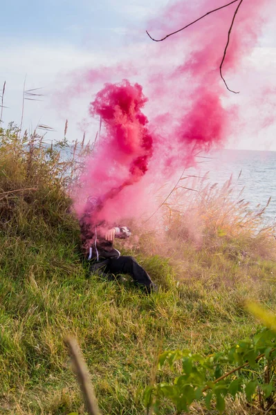 A boy in a black sports suit sits on the green grass, the boy has a crimson smoke bomb in his hand and it smokes into the sky.