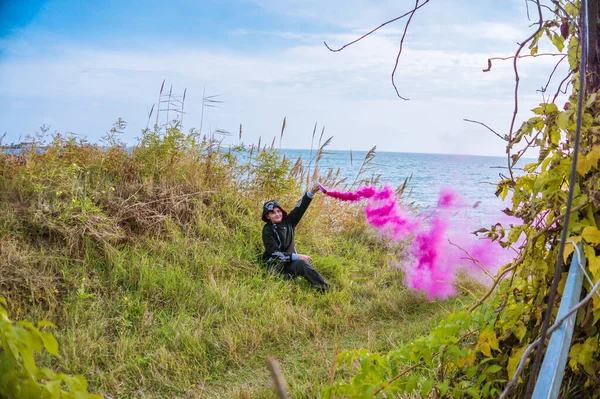 A boy in a black sports suit sits on the green grass, the boy has a crimson smoke bomb in his hand and it smokes into the sky.