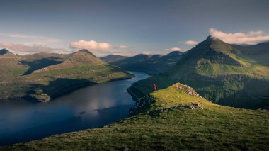 Faroe Adaları 'ndaki Gjogv kasabası yakınlarındaki Hvithamar' da kırmızı ceketli bir adam, Eysturoy adası fiyort panoramik manzaralı, mavi gökyüzü