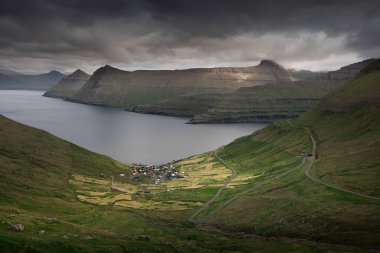 Faroe Adaları 'nın panoramik dağ manzarası, Eysturoy adasının kıyısında Funningur köyü, bulutlu ve okyanuslu dramatik gökyüzü, yeşil vadiyi saran dağ geçidi.