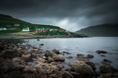 Faroe Adaları 'ndaki Eysturoy köyündeki Elduvk köyünde renkli evleri ve kilisesi olan körfez; ön planda kayalar ve taşlar, gökyüzünde dramatik kara bulutlar olan okyanus