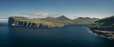 Gündüz vakti Faroe Adaları 'ndaki Risin og Kellingin kaya oluşumlarıyla Eysturoy adasının sahil şeridinde panoramik manzara. Güneş ve mavi gökyüzü, mavi okyanus ve yeşil uçurumlar.