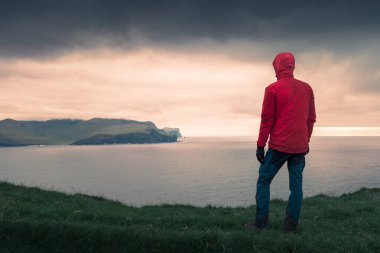 man in red jacket looking out over cliffs at Eysturoy in sunset, Faroe Islands clipart