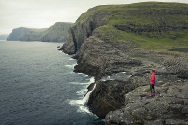 Sahilde mor ceketli bir kadın, Faroe Adaları 'ndaki Leitisvatn Gölü' ndeki Vagar adasında şelale ve uçurumlar.
