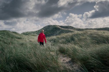 Kum tepelerinde kırmızı ceketli bir adam, Danimarka sahillerinde yeşil çimenler, dramatik gökyüzünde bulutlar.