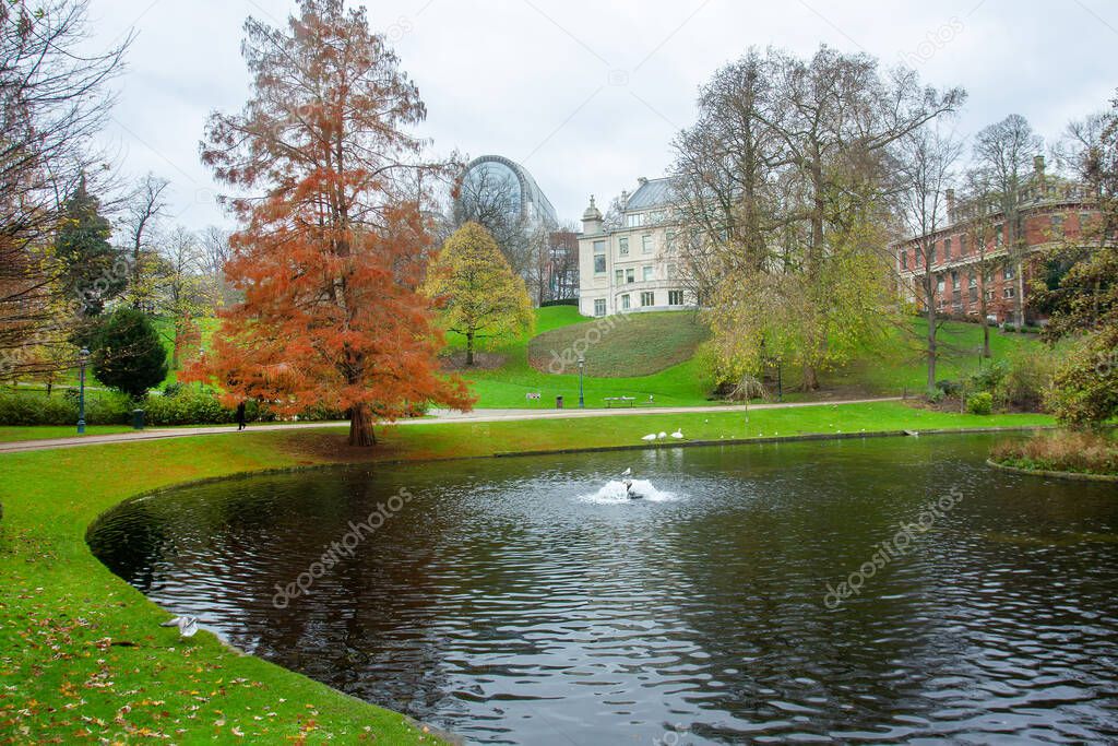 Pretty park with pond , and trees and pathes and EU building at far distance . Small fountain in pond.. White swans and unrecognizable person walking.