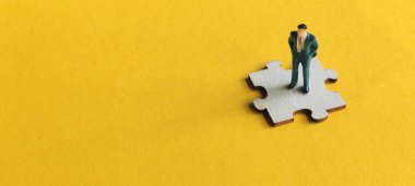 Tek bir yapboz parçasının üzerinde duran minyatür bir işadamı, sarı arka plandaki önemli çözümü, liderliği ve bir projenin ya da sistemin eksik parçasını sembolize ediyor..