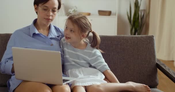 Lycklig mamma och söt liten dotter som har roligt sittandes på soffan hemma pratar med hjälp av att lära sig bärbar dator, leende mamma undervisar liten flicka på nätet, gör webcam videosamtal tittar på datorskärmen — Stockvideo