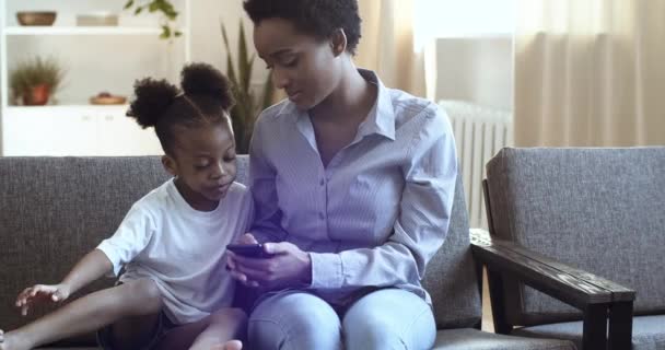 Kędzierzawa Afroamerykanka i mała dziewczynka siedzą na domowej kanapie. Kobieta pokazuje coś małej uroczej córce na ekranie smartfona. Mama z małym dzieckiem podsłuchiwanie, przewijanie gry na telefon komórkowy — Wideo stockowe