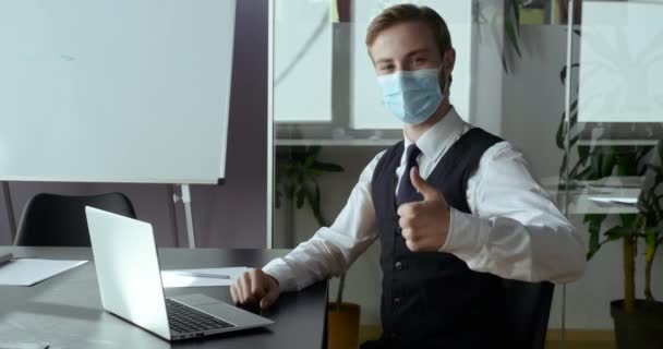 Молодий студент успішний бізнесмен носить захисну медичну маску на чоловічому обличчі, бізнесмен сидить в офісі показує знак схвалення та підтримки на камеру рукою, ставить великий палець як символ — стокове відео