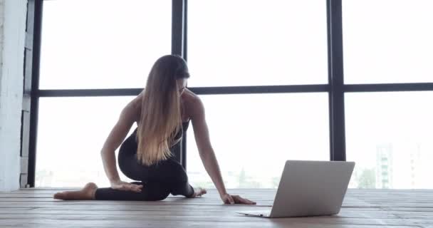Bela menina do esporte caucasiano feminino sentado no chão em casa em frente ao laptop assistindo exercícios de vídeo. Mulher dançarina treinador de fitness fazendo alongamento olhando na tela do computador, lição on-line — Vídeo de Stock