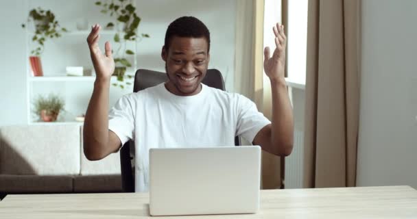 Bel homme afro-américain célébrant le succès alors qu'il était assis à la maison au bureau avec un ordinateur portable. Noir confiant étudiant porte casual t-shirt blanc regarde écran d'ordinateur surpris se sent triomphe — Video