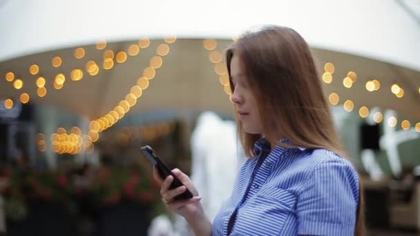 Close-up view of beautiful young girl in striped shirt watching video on smartphone then looking at camera and laughing sweetly on defocused background of fountain and lights of restaurant on street. — Stock Video