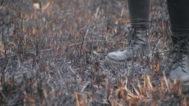 Shot from below in slow motion of persons legs in leather stylish youth boots standing on the burnt grass after serious fire in field. Man stomps on burned earth, ashes fly up in air from the wind. — Stock Video