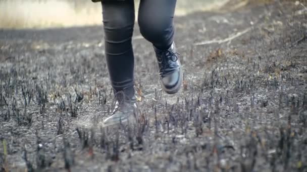 Movendo a vista frontal do campo deserto correndo ao longo da grama cinza queimada após o fogo da menina em botas de couro preto na moda com solas altas e leggings em tempo de primavera ensolarado, em câmera lenta. — Vídeo de Stock