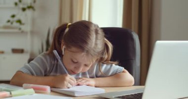 Karantinada uzaktan öğrenme. Küçük anaokulu öğrencisi dizüstü bilgisayarla uzaktan kumanda okuyup not tutuyor. Kulaklık takan çocuk okulu öğrencisi internet videosu dersi izliyor.