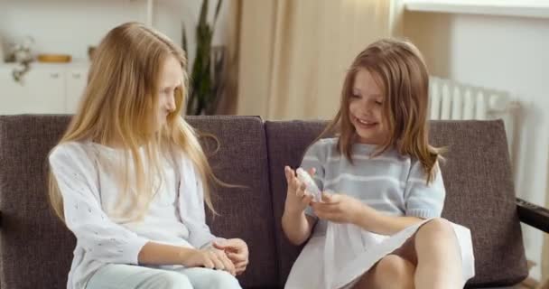 Due piccole ragazze bionde coppia di sorelle amiche sedute a casa sul divano insieme durante la quarantena utilizzando una bottiglia di gel alcolico liquido per trattare le mani, i bambini che utilizzano igienizzanti contro i batteri del virus — Video Stock