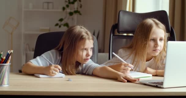 2人の幸せなかわいい小さな友達の女の子が自宅のテーブルやクラスで座って、ノートパソコンで遠隔研究し、宿題をします。論理学の練習、 eラーニングのホームスクールの概念を議論する笑顔の小さな姉妹 — ストック動画