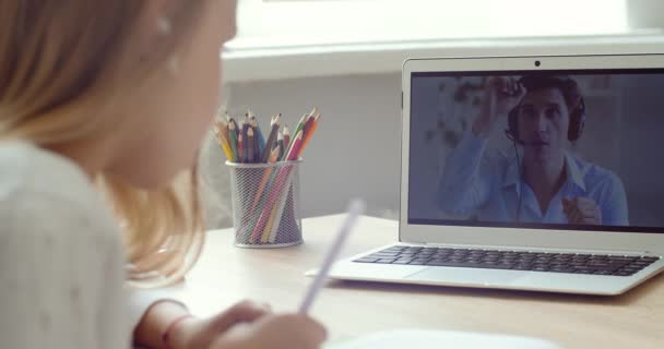 Mądra dziewczynka używa laptopa do rozmowy wideo z nauczycielem online. Ekran pokazuje wideo wykład z korepetytorem wyjaśniający temat z klasy lub domu, uczennica zapisuje informacje, e-edukacja — Wideo stockowe