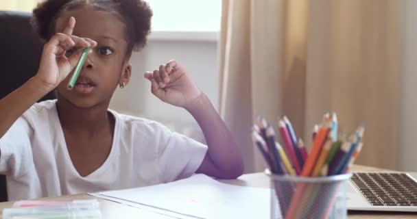 Kis aranyos afrikai lány göndör haj ül az asztalnál otthon, játszik egyedül, felhívja a papírt, választ színes ceruza, részt vesz a kreativitás tanulmányok távolról, észrevételek a kép, otthon tanulás koncepció — Stock videók