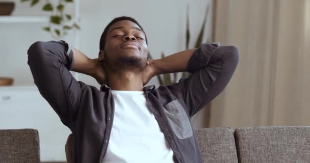 Retrato de hombre africano joven estudiante sentado en el sofá en casa en la habitación interior con los brazos cruzados detrás de la cabeza masculina cerrado los ojos soñando con planes futuros pensando descansando relajado en el descanso — Vídeo de stock